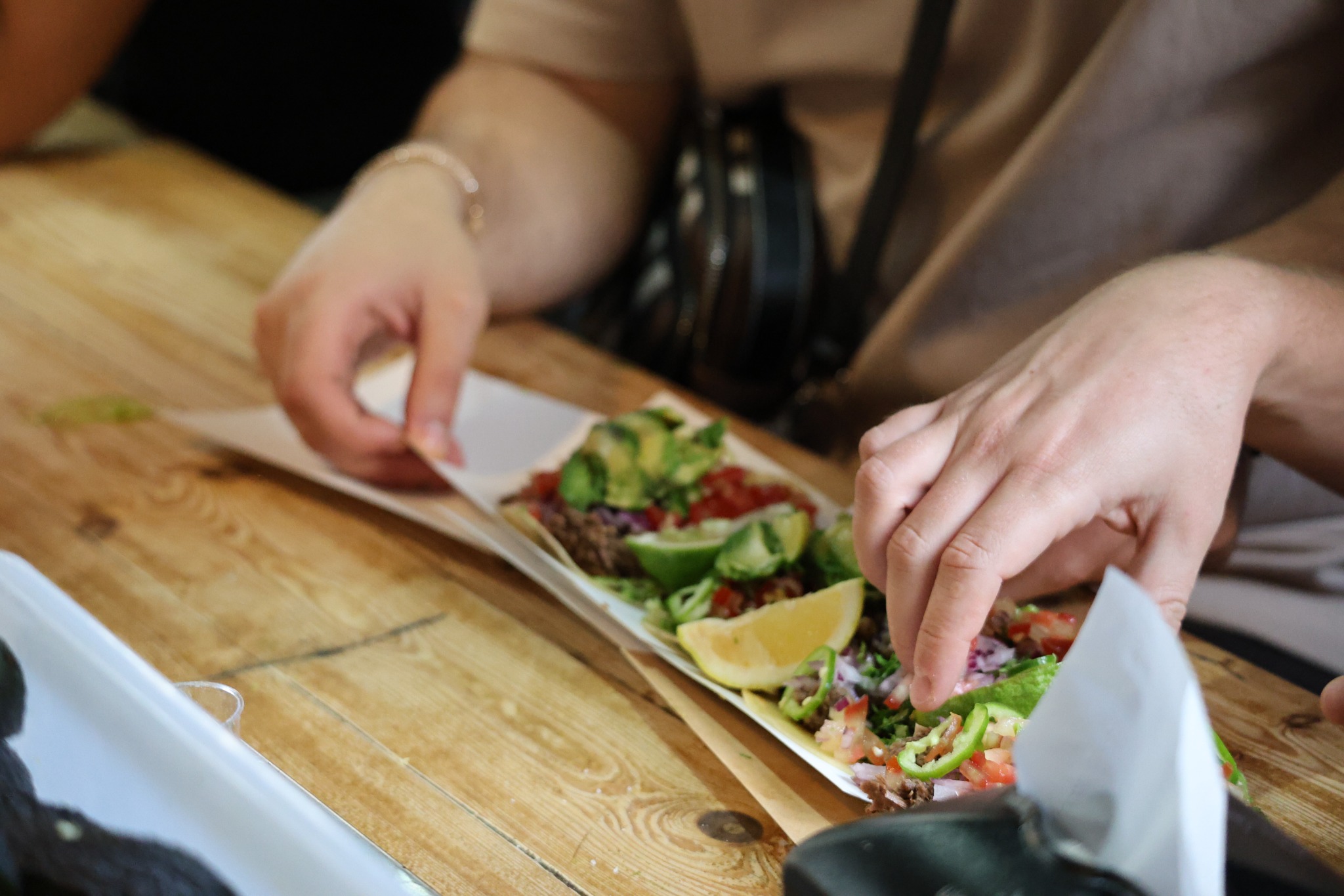 A diner reaches toward tacos topped with avocado, diced tomato, red onion, herbs and green pepper rings on a wooden counter, with citrus wedges alongside.