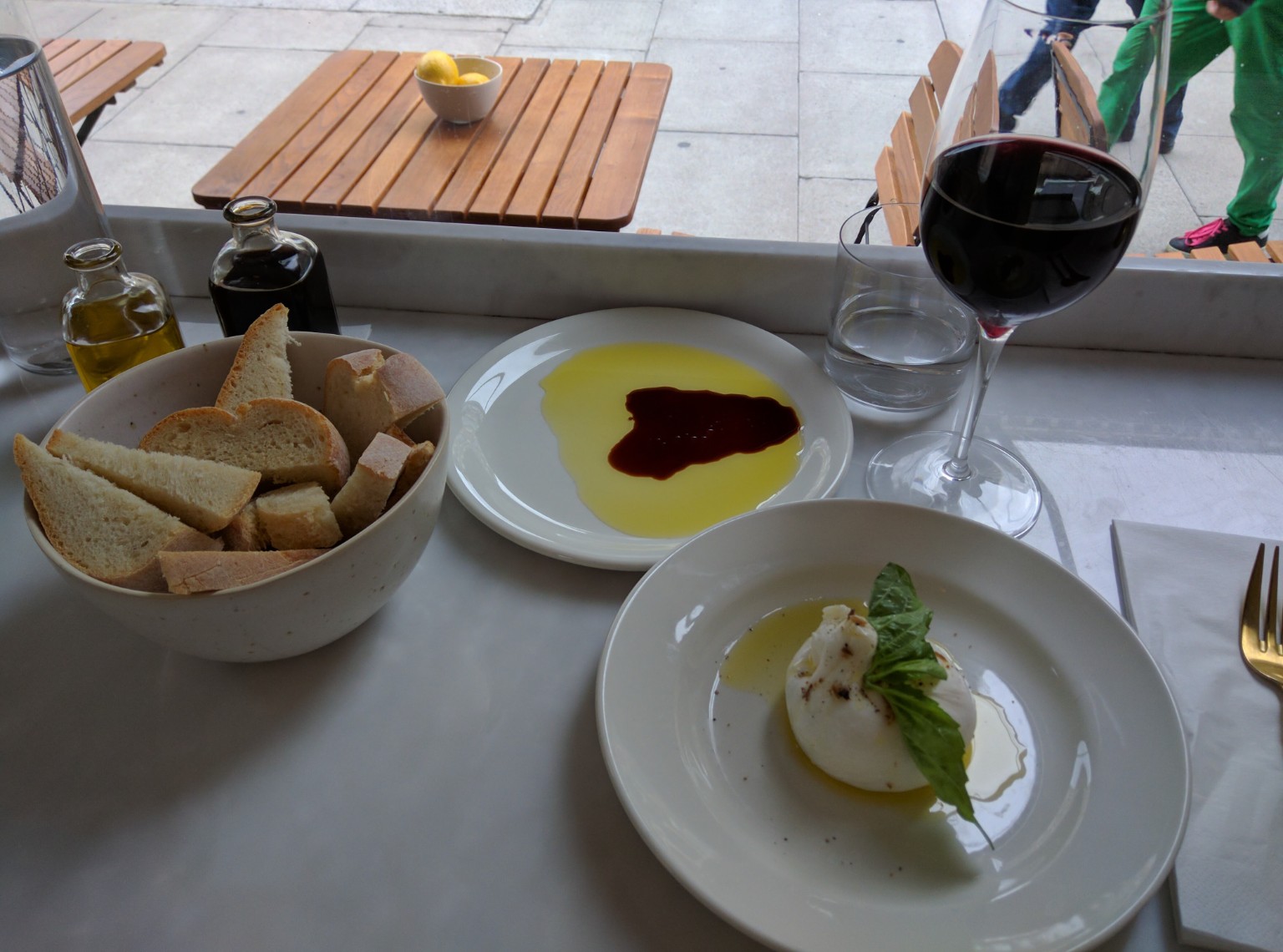 Burrata topped with basil and olive oil beside crusty bread, a plate of oil and dark vinegar, and a glass of red wine at a window counter.