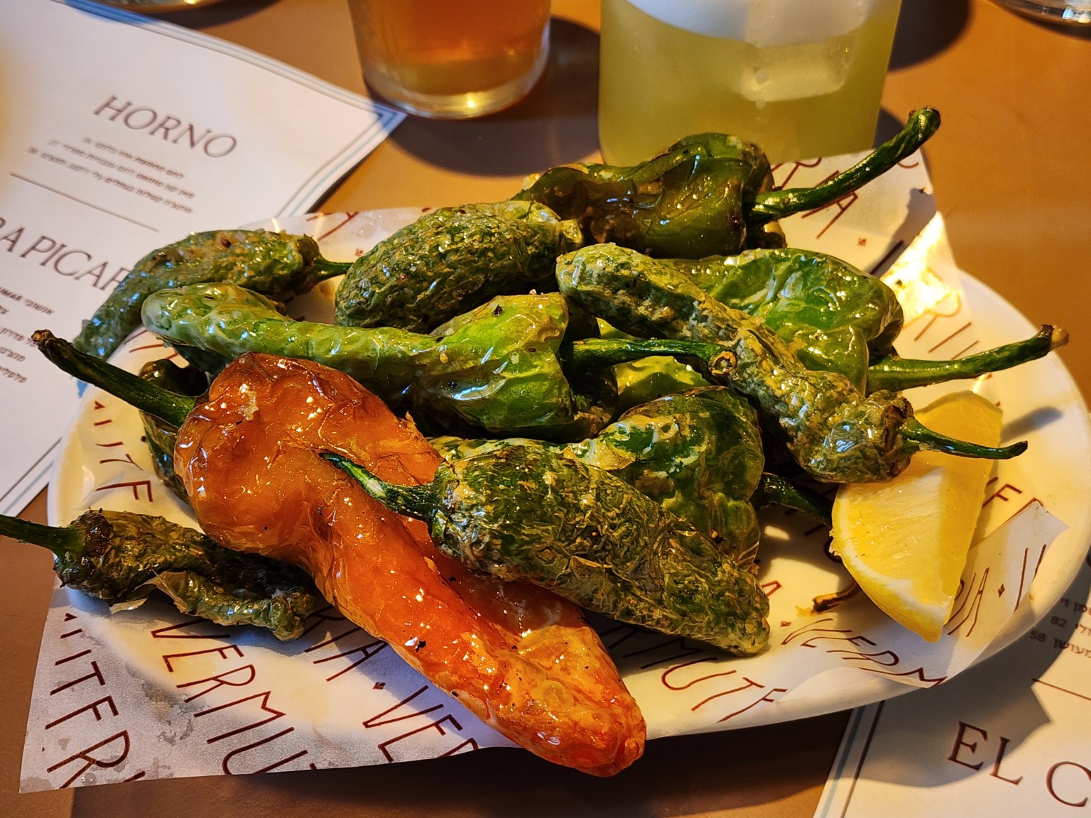 A plate of blistered Padrón peppers, including one orange-red pepper, with lemon wedges at a tapas restaurant