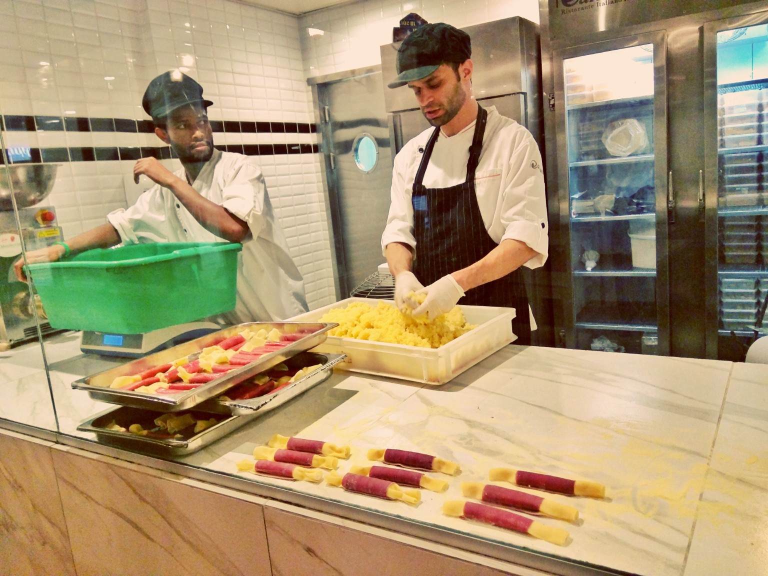 Two cooks making pasta behind glass, with pink and yellow candy-shaped parcels on a flour-dusted counter.