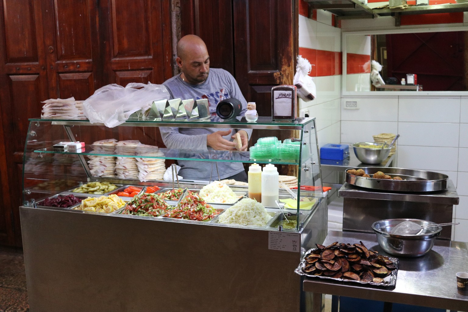 Vendor preparing pita behind a glass counter filled with chopped salads, pickles and cabbage, beside falafel and a tray of fried eggplant slices.
